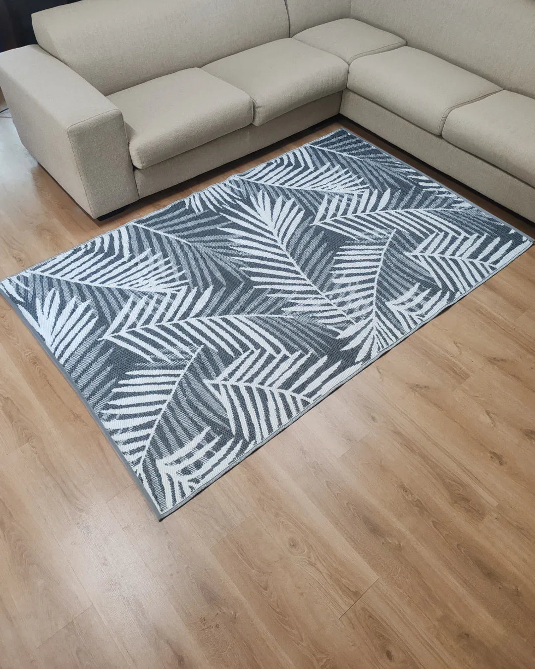 A modern living room with a beige sectional sofa and the Florastyle By Hingham Medium Hanoi Outdoor Reversible Mat—lightweight, double woven, water resistant—featuring a tropical leaf pattern on a light wood floor.