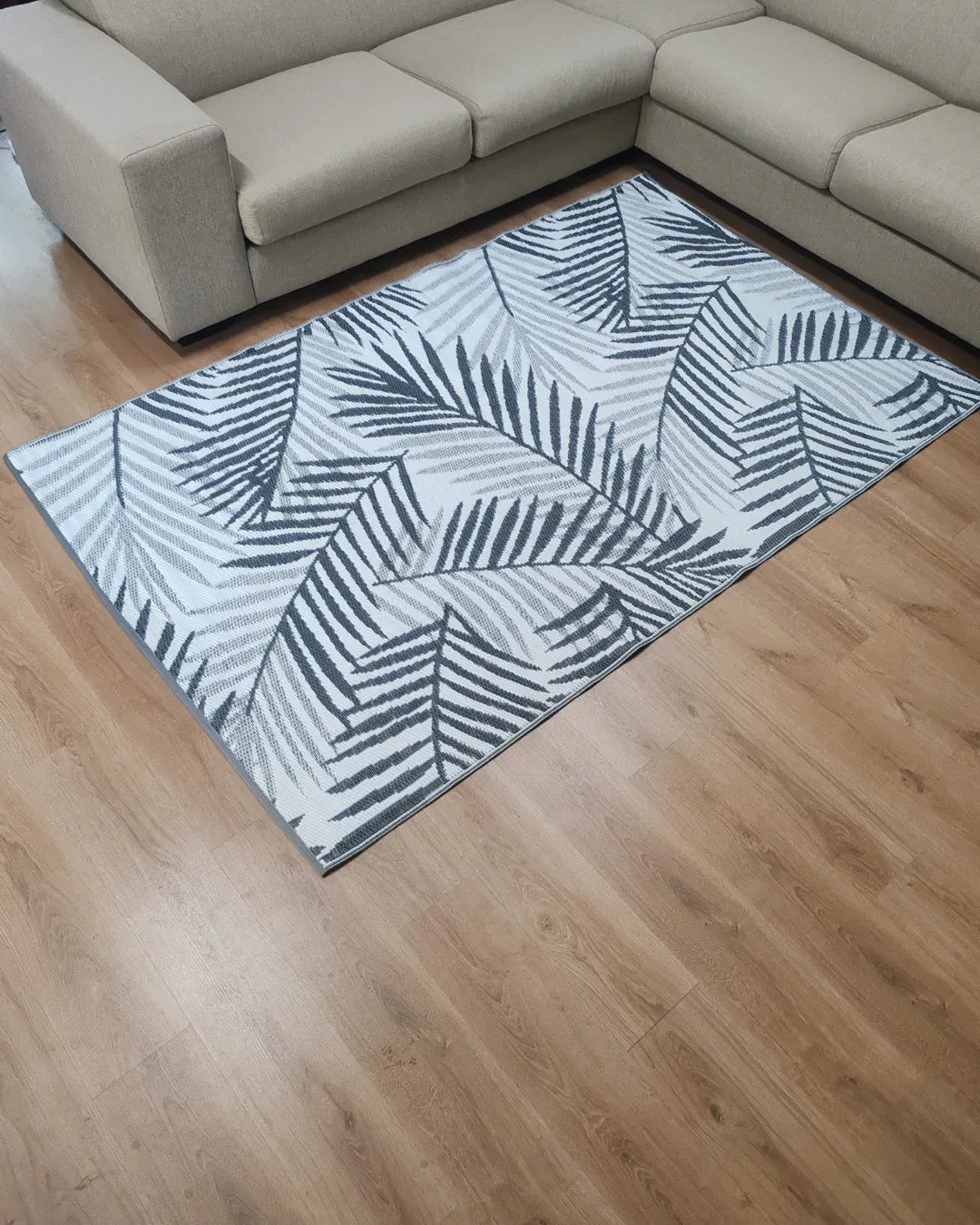 The Medium Hanoi Outdoor Reversible Mat by Florastyle By Hingham, a lightweight, double woven rug with a black and white palm leaf design, is placed on a light wooden floor in front of a beige sectional sofa in the living room.