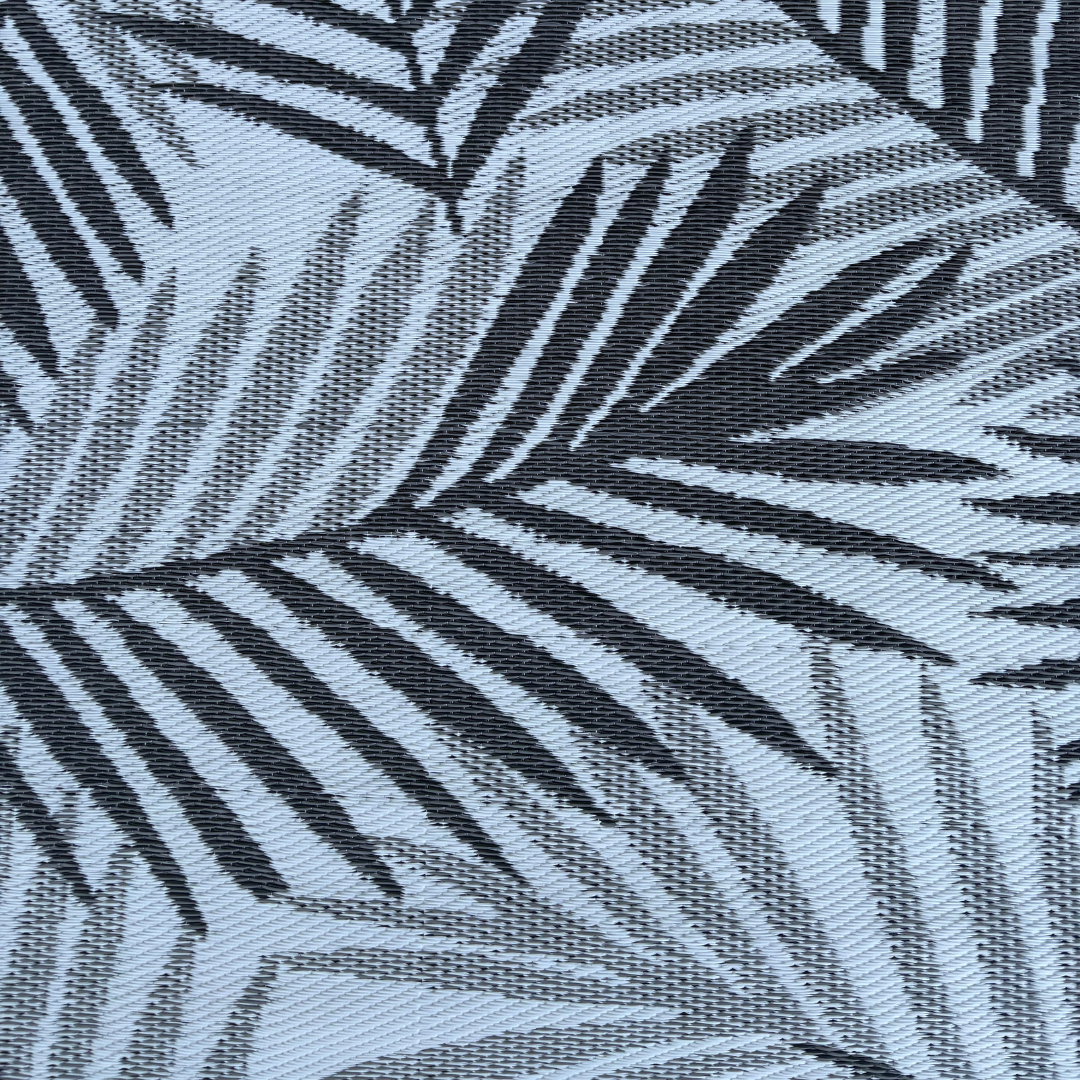 Black palm leaf silhouettes overlap on a light blue background, creating a bold, tropical look. The Medium Hanoi Outdoor Reversible Mat by Florastyle By Hingham is lightweight, double woven, and water resistant for comfort and durability.