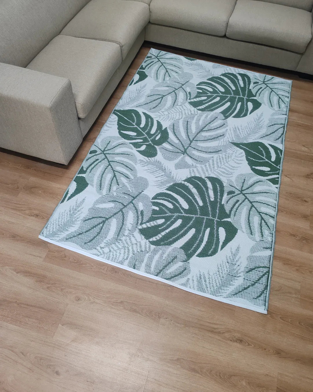 The Florastyle By Hingham Medium Lima Outdoor Reversible Mat, a lightweight, double-woven, water-resistant rug with a green and white tropical leaf pattern, is placed on a light wood floor and surrounded by a beige sectional sofa.