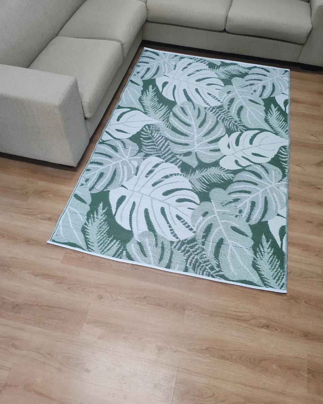 The Florastyle By Hingham Medium Lima Outdoor Reversible Mat, a lightweight, water-resistant rug with a green and white tropical leaf design, is placed on a light wooden floor between two beige sectional sofas.