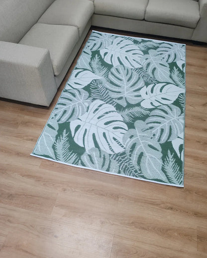 The Florastyle By Hingham Medium Lima Outdoor Reversible Mat, a lightweight, water-resistant rug with a green and white tropical leaf design, is placed on a light wooden floor between two beige sectional sofas.