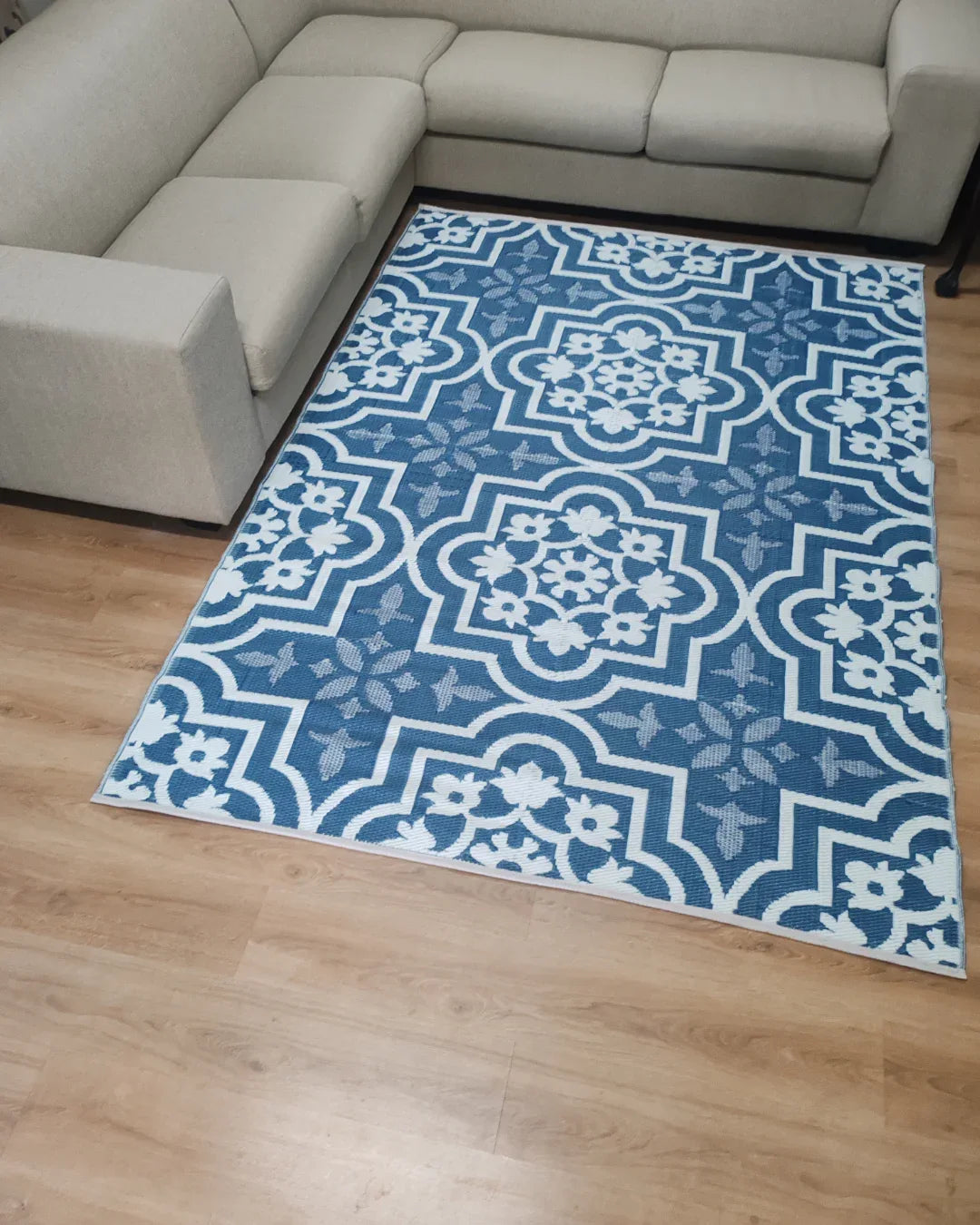 The Large Marrakesh Outdoor Reversible Mat by Florastyle By Hingham, a lightweight blue and white geometric and floral rug, is placed on a light wood floor beside a beige sectional sofa in the living room.