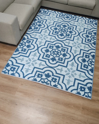 The Florastyle By Hingham Large Marrakesh Outdoor Reversible Mat, a lightweight, double-woven, water-resistant rug with blue and white floral and geometric patterns, is placed on a light wood floor beside a beige sectional sofa.