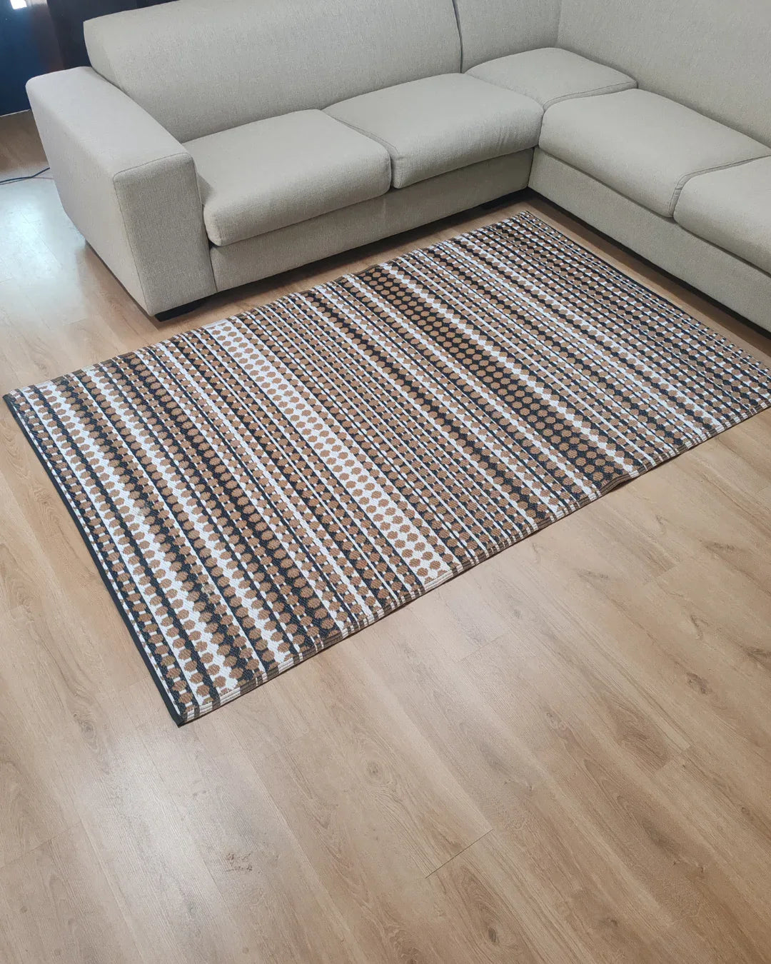 A lightweight beige sectional sofa sits on a light wood floor next to the Florastyle By Hingham Medium Savannah Outdoor Reversible Mat, featuring a geometric pattern in brown, beige, and white.
