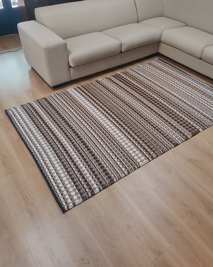 The Florastyle By Hingham Medium Savannah Outdoor Reversible Mat, a lightweight, double woven rug in a striped brown, black, and white pattern, sits on a light wood floor in front of a beige sectional sofa.