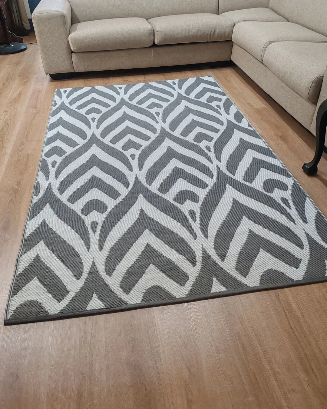 A modern living room with a light wood floor, beige sectional sofas, and the Florastyle By Hingham Medium Sienna Outdoor Reversible Mat—lightweight, double woven, water-resistant with a bold gray and white geometric pattern.