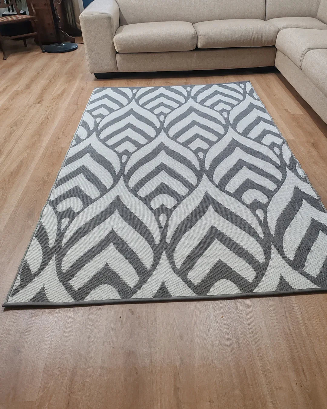 A Medium Sienna Outdoor Reversible Mat by Florastyle By Hingham, lightweight and water-resistant with a double woven geometric leaf pattern, is placed on a light wood floor in front of a beige sectional sofa in the living room.