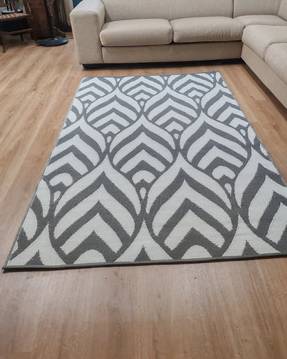 A Medium Sienna Outdoor Reversible Mat by Florastyle By Hingham, lightweight and water-resistant with a double woven geometric leaf pattern, is placed on a light wood floor in front of a beige sectional sofa in the living room.