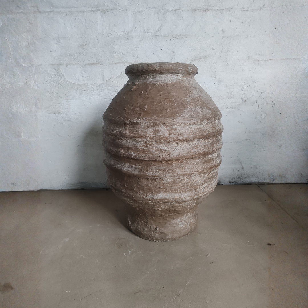 The Sparta Organic Fiberglass Pot - Earth Range by Florastyle By Hingham, featuring a wide body, narrow neck, and horizontal ridges, sits on a plain floor against a white textured wall.