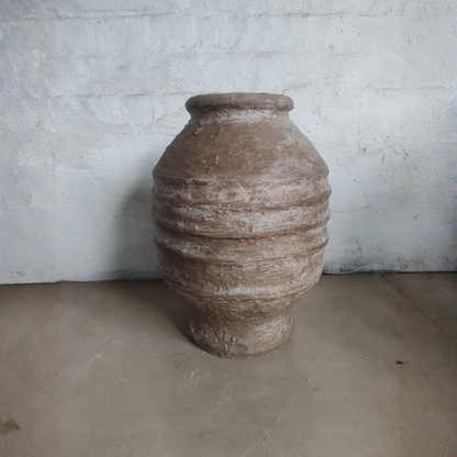 The Sparta Organic Fiberglass Pot - Earth Range by Florastyle By Hingham, featuring a wide body, narrow neck, and horizontal ridges, sits on a plain floor against a white textured wall.