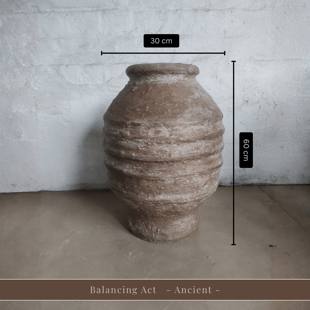 A spacious Sparta Organic Fiberglass Pot from Florastyle By Hingham, earth-toned and lightweight, rests against a white wall. Dimensions: 30 cm wide by 60 cm tall. Text below reads, Balancing Act - Ancient -.
