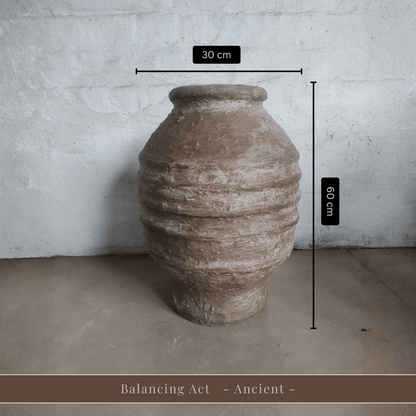 A spacious Sparta Organic Fiberglass Pot from Florastyle By Hingham, earth-toned and lightweight, rests against a white wall. Dimensions: 30 cm wide by 60 cm tall. Text below reads, Balancing Act - Ancient -.