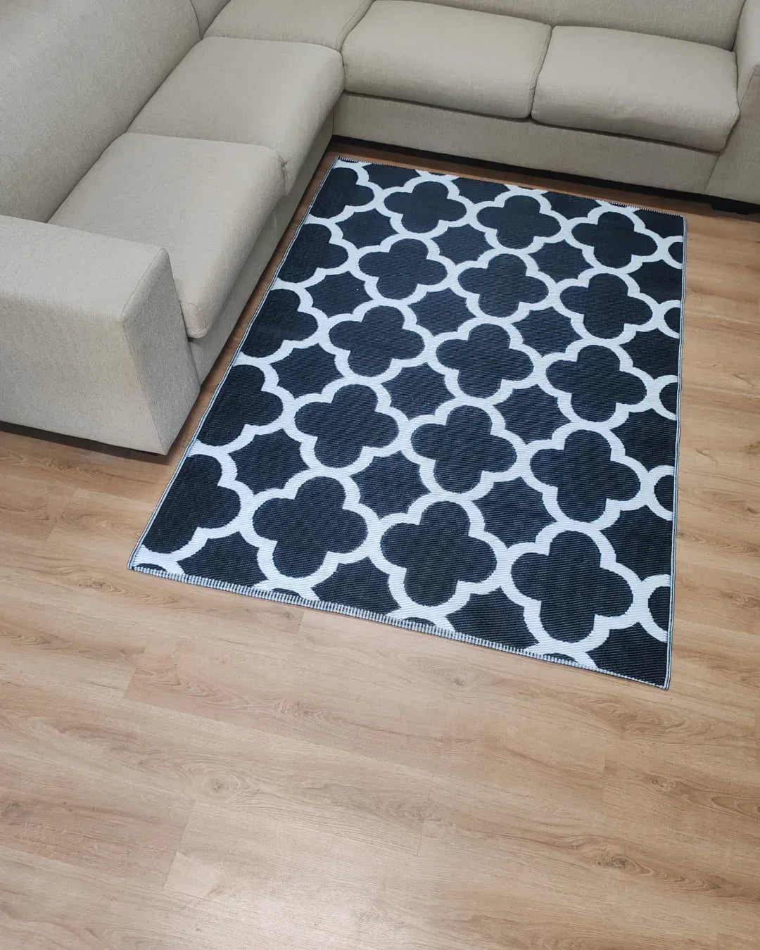 A beige, lightweight sectional sofa surrounds the Florastyle By Hingham Medium Tripoli Outdoor Reversible Mat, featuring a blue and white geometric quatrefoil pattern, set on a light wood living room floor.
