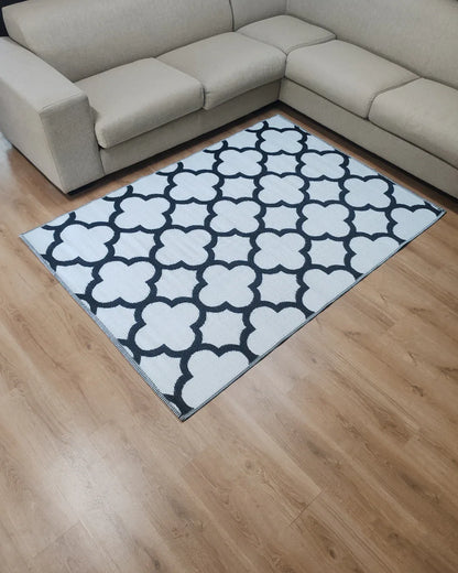 A light beige sectional sofa surrounds the Florastyle By Hingham Medium Tripoli Outdoor Reversible Mat—a lightweight, double-woven rug with a bold black geometric quatrefoil pattern—set on a light wooden floor.