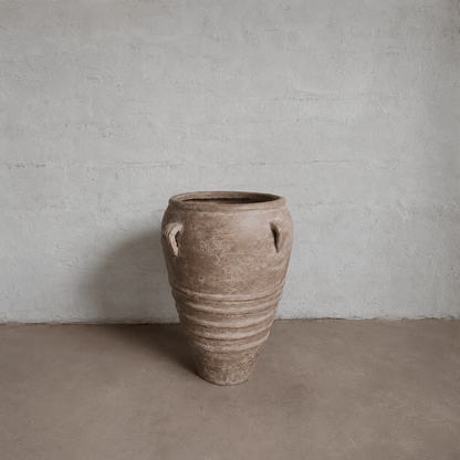 The Athens Organic Fiberglass Pot - Earth Range by Florastyle By Hingham, featuring horizontal ridges, small handles, and an earthy, weathered look, stands on a bare floor against a textured light plaster wall.
