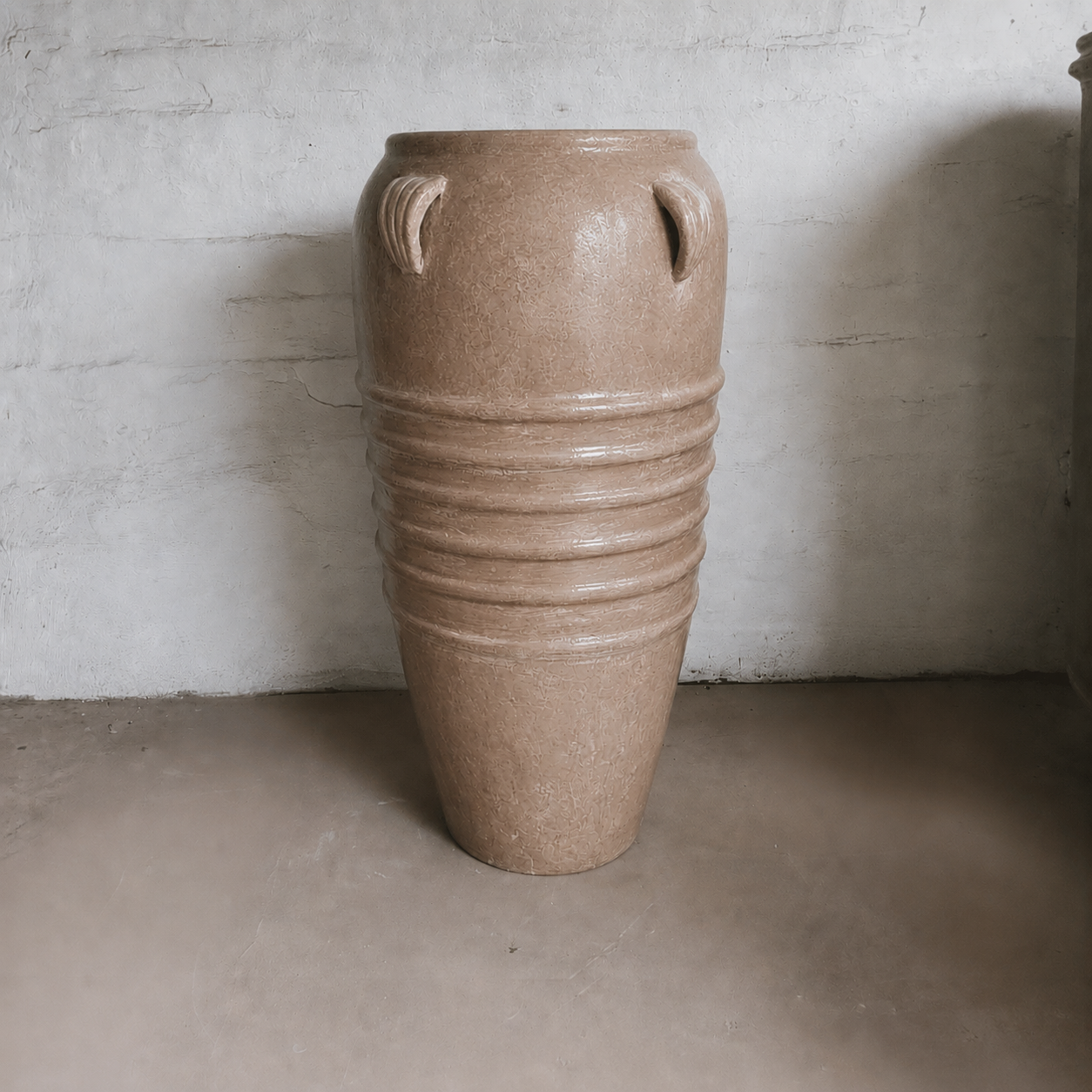 The Athens Organic Fiberglass Pot - Earth Range by Florastyle By Hingham features a tall, brown finish with ribbed detailing and twin handles, evoking ancient Greek pottery style, displayed against a white wall on a concrete floor.