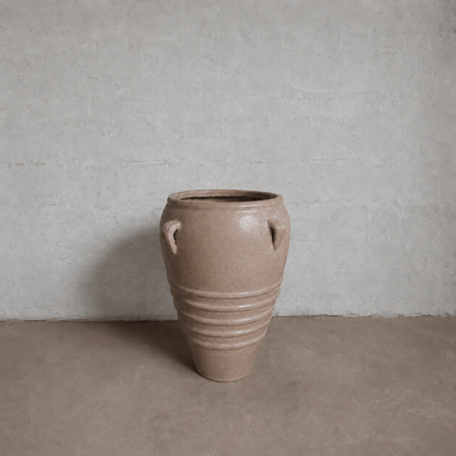 The Florastyle By Hingham Athens Organic Fiberglass Pot - Earth Range is a large tan vase inspired by ancient Greek pottery, with a narrow base, wide rim, two top handles, and lower half ridges, set against a light gray wall and brown floor.