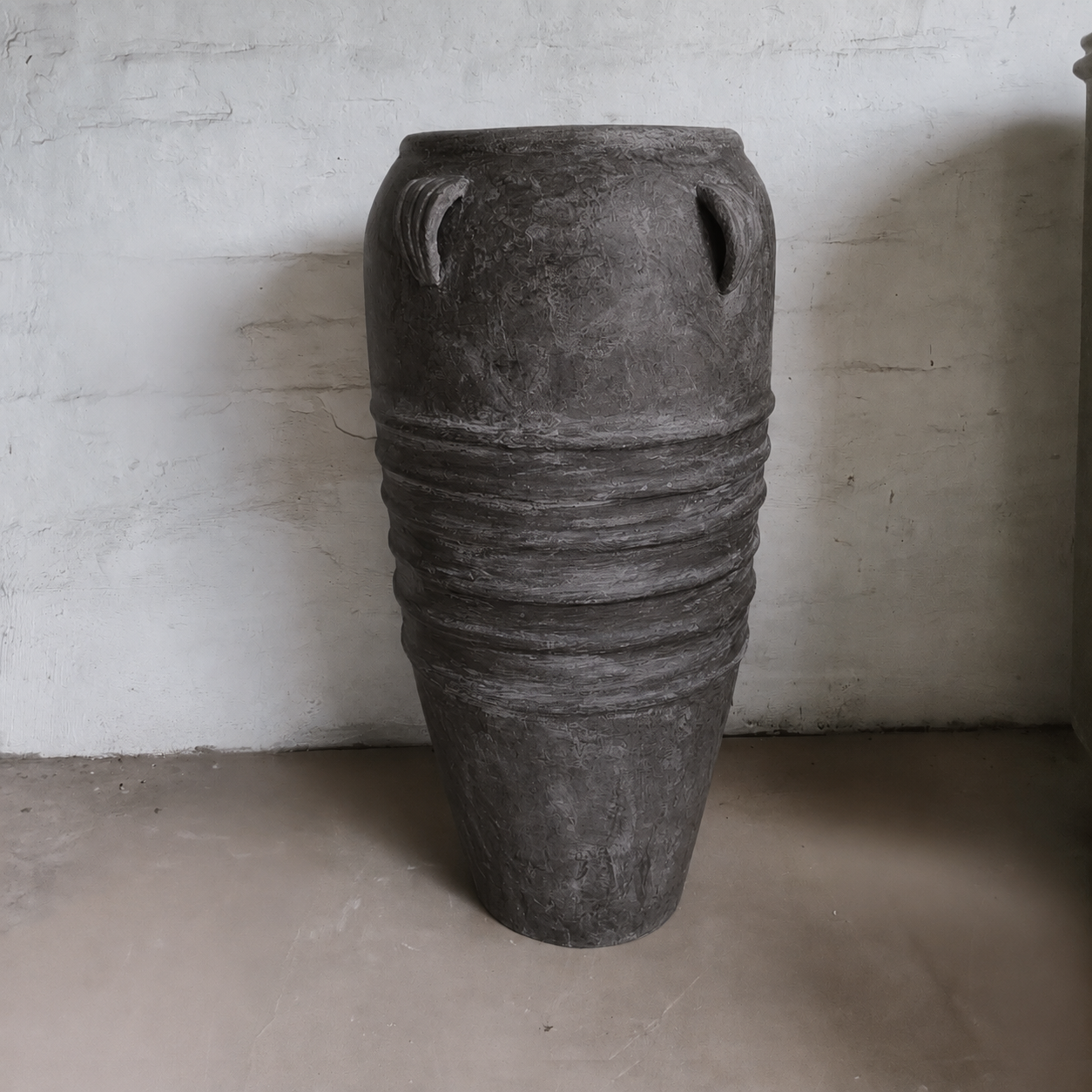 The Athens Organic Fiberglass Pot – Earth Range by Florastyle By Hingham, featuring a wide body, narrow base, and decorative ridges, stands against a white wall on a cement floor, echoing ancient Greek pottery style.