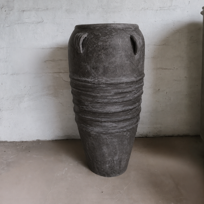 The Athens Organic Fiberglass Pot – Earth Range by Florastyle By Hingham, featuring a wide body, narrow base, and decorative ridges, stands against a white wall on a cement floor, echoing ancient Greek pottery style.