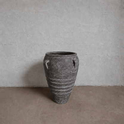 The Athens Organic Fiberglass Pot – Earth Range by Florastyle By Hingham, featuring two small handles and horizontal ridges, sits on a brown floor against a textured light gray wall, echoing ancient Greek pottery.