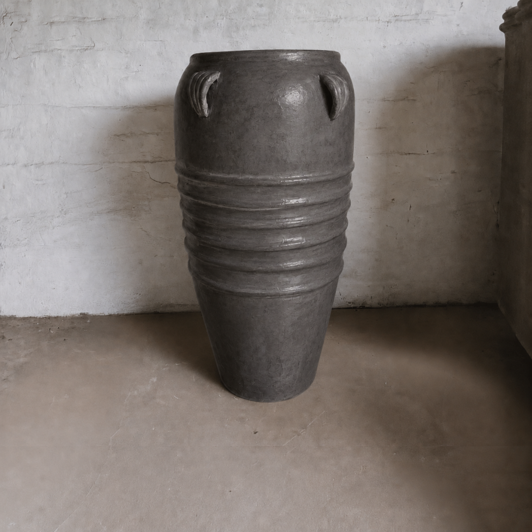 The Athens Organic Fiberglass Pot - Earth Range by Florastyle By Hingham features a large, dark finish with four top handles and horizontal ridges, evoking ancient Greek style. It stands on a concrete floor against a white brick wall.