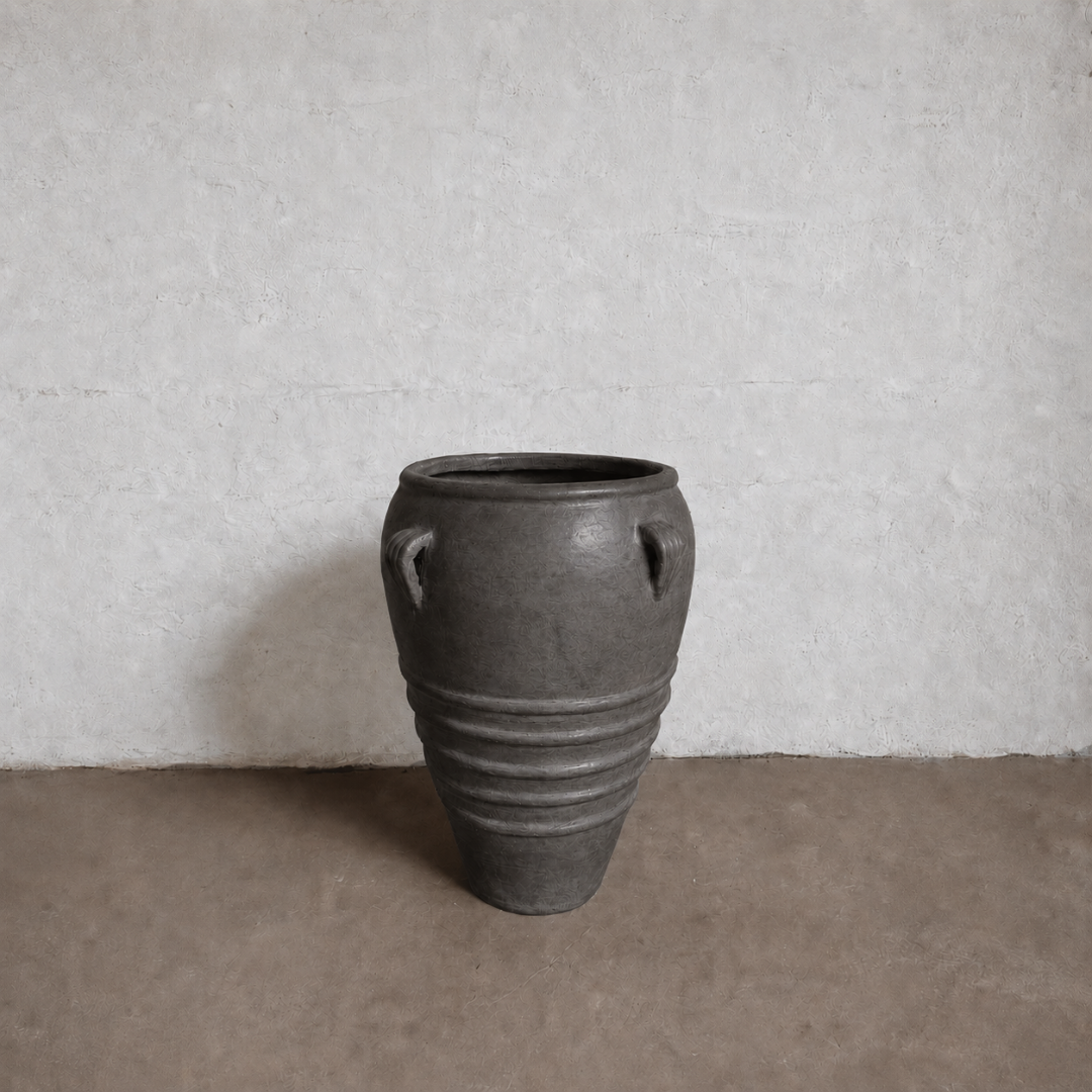 The Athens Organic Fiberglass Pot - Earth Range by Florastyle By Hingham, a large dark gray vase with two small handles and horizontal ridges, sits on a brown floor against a textured white wall.