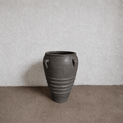 The Athens Organic Fiberglass Pot - Earth Range by Florastyle By Hingham, a large dark gray vase with two small handles and horizontal ridges, sits on a brown floor against a textured white wall.