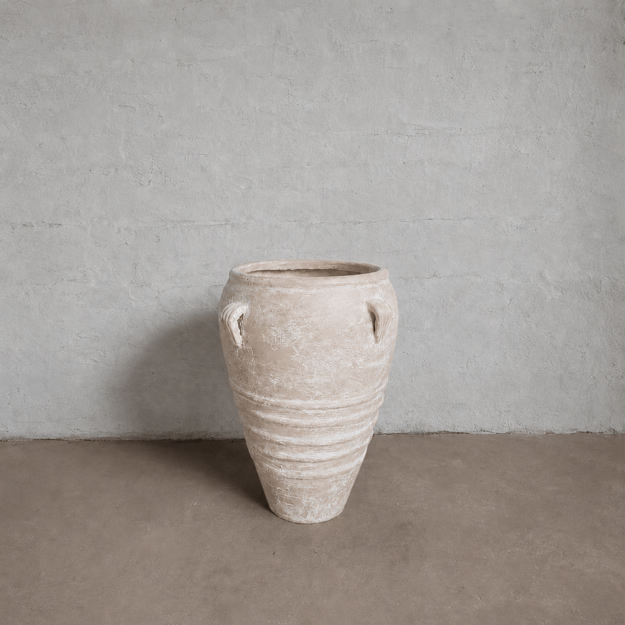 The Athens Organic Fiberglass Pot - Earth Range by Florastyle By Hingham, featuring two small handles and a weathered finish, stands on a plain brown floor against a light gray textured wall.