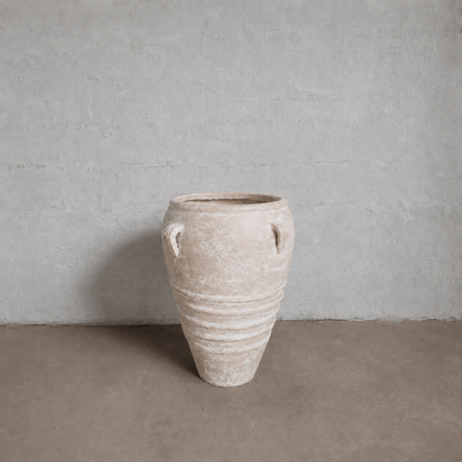 The Athens Organic Fiberglass Pot - Earth Range by Florastyle By Hingham, featuring two small handles and a weathered finish, stands on a plain brown floor against a light gray textured wall.