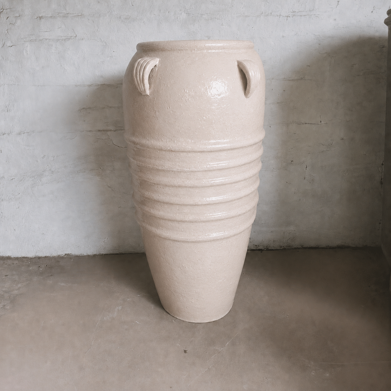The Athens Organic Fiberglass Pot from Florastyle By Hingham’s Earth Range, featuring vertical handles and horizontal ridges inspired by ancient Greek pottery, is displayed against a light textured wall on a concrete floor.