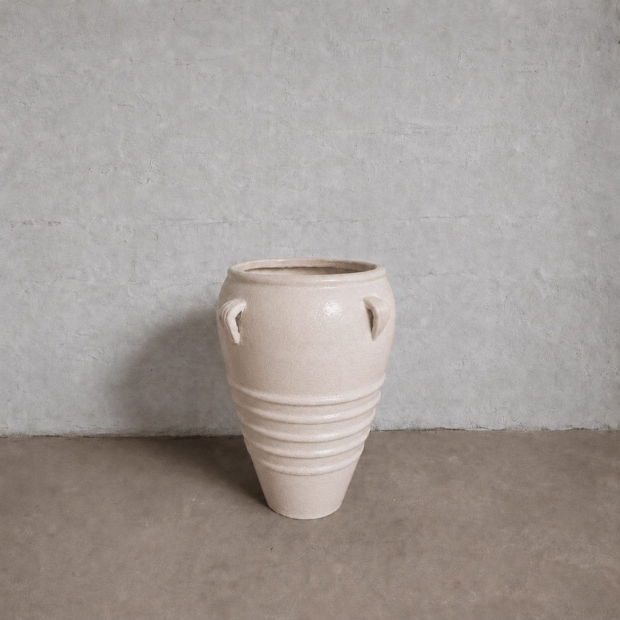 The Athens Organic Fiberglass Pot - Earth Range by Florastyle By Hingham, with two small handles and ridged base details, stands against a minimalist gray wall on a smooth floor.