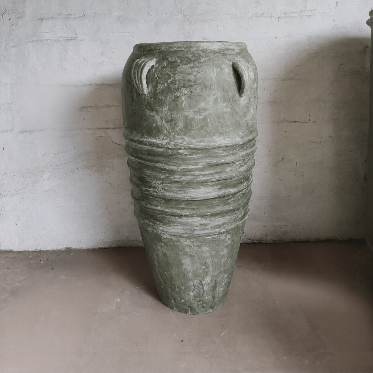 The Athens Organic Fiberglass Pot from Florastyle By Hingham features horizontal ridges and rounded handles, offering timeless Greek-inspired style in an earth-toned finish—perfect against a white textured wall.