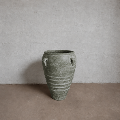 The Athens Organic Fiberglass Pot from Florastyle By Hingham’s Earth Range, with a textured greenish-gray finish and two small handles, sits on a brown floor against a plain light gray wall, evoking ancient Greek pottery.
