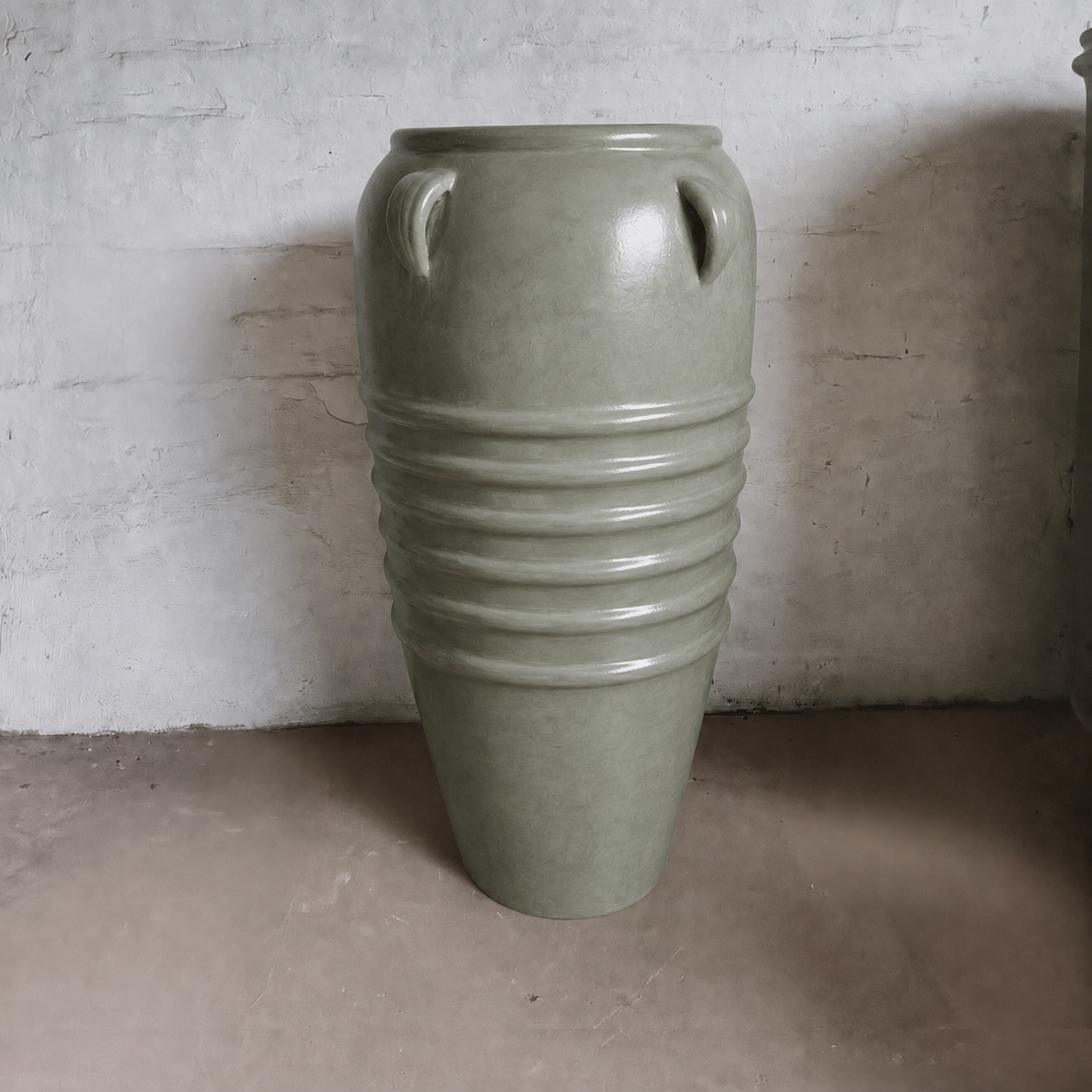 The Athens Organic Fiberglass Pot - Earth Range by Florastyle By Hingham, featuring a tall green form with four upper handles and horizontal ridges, sits on a concrete floor against a textured white wall.