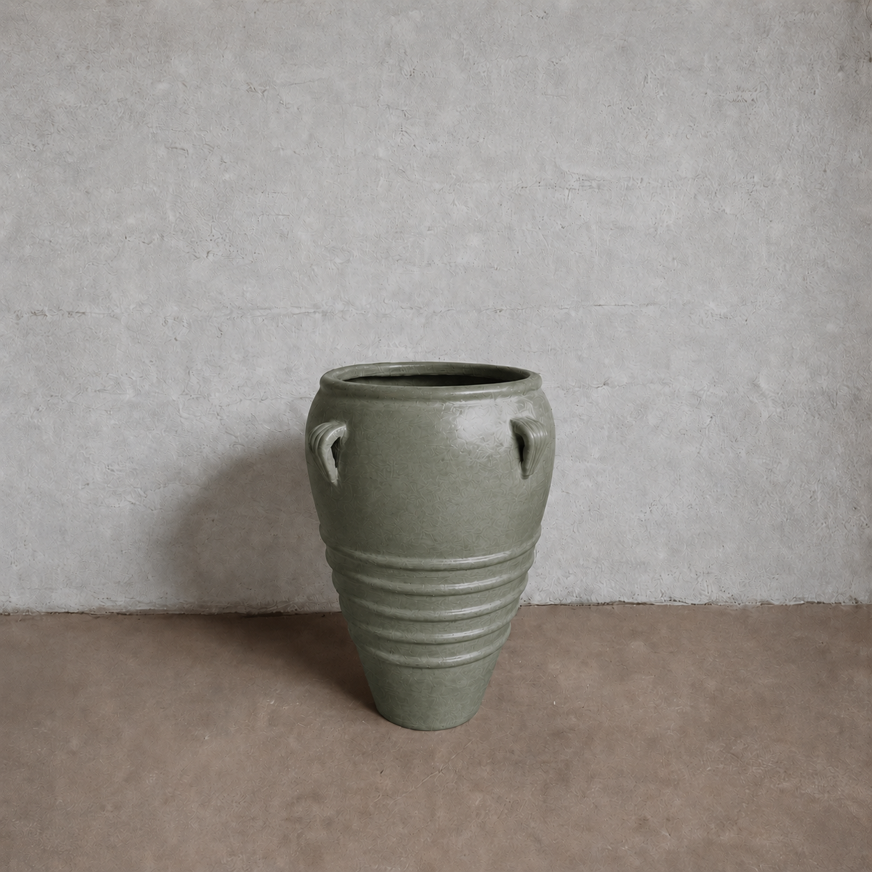 The Athens Organic Fiberglass Pot - Earth Range by Florastyle By Hingham, a large green vessel with twin top handles and horizontal ridges, evokes ancient Greek pottery and sits on a brown floor against a textured light gray wall.