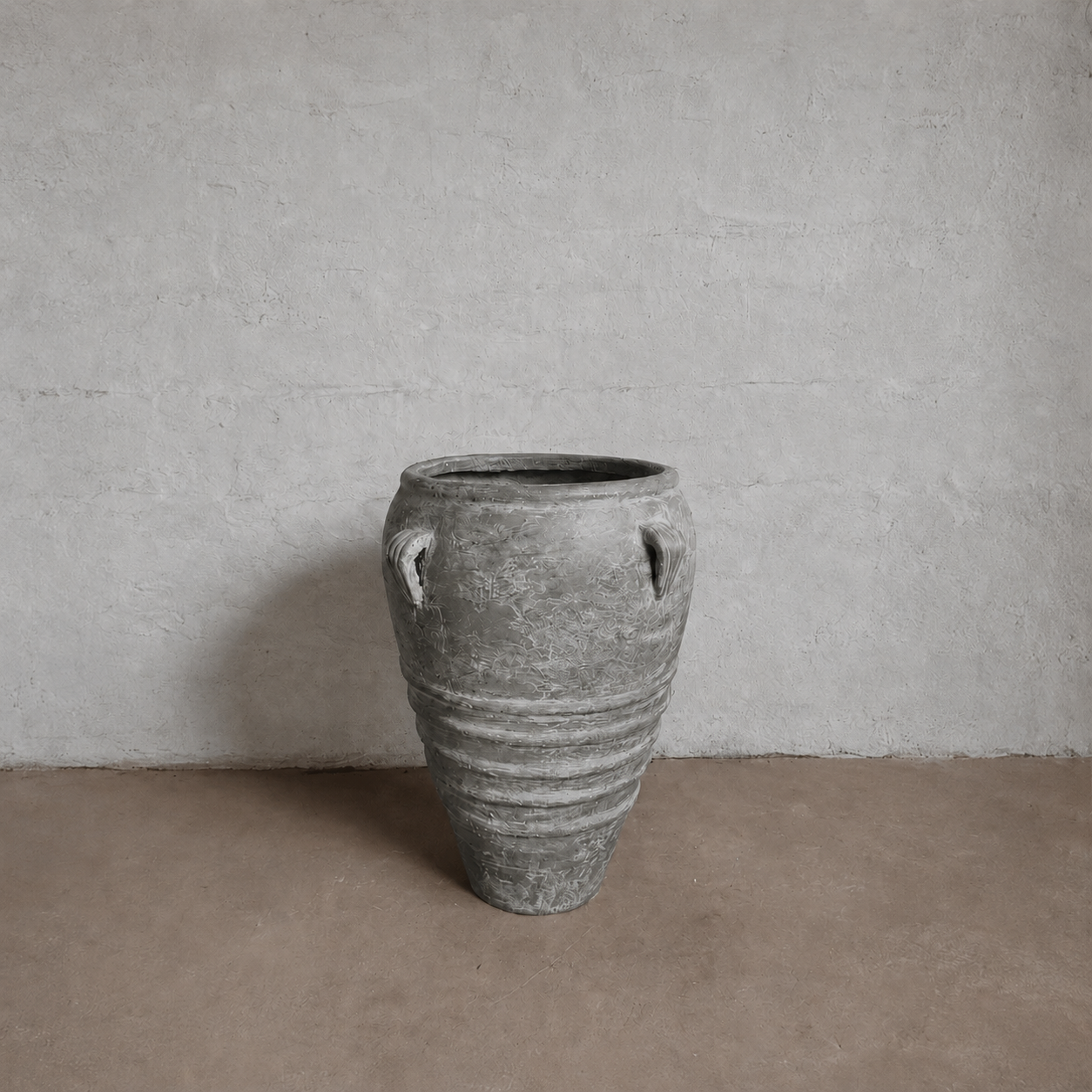 The Athens Organic Fiberglass Pot - Earth Range by Florastyle By Hingham, with its weathered gray finish and two side handles, stands on a brown floor against a textured light gray wall, evoking the elegance of ancient Greek pottery.