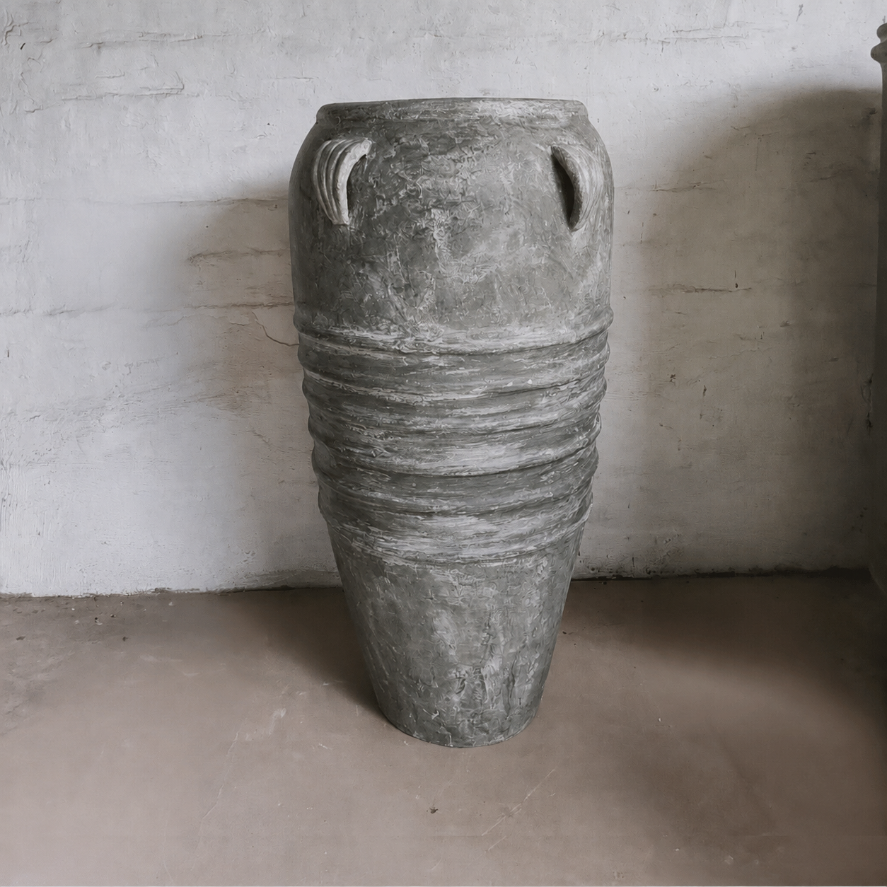 The Florastyle By Hingham Athens Organic Fiberglass Pot - Earth Range, a large gray planter with dual handles and midsection ridges, stands on a concrete floor against a textured white wall.