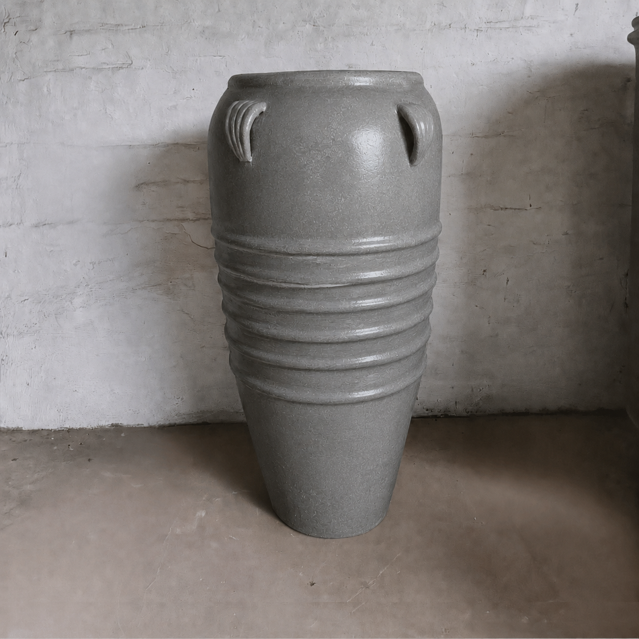 The Athens Organic Fiberglass Pot - Earth Range by Florastyle By Hingham, featuring a tall gray form with four small rim handles and midsection ridges, stands on a light floor against a textured white wall.