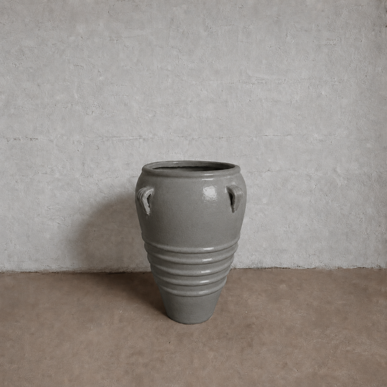 The Athens Organic Fiberglass Pot - Earth Range by Florastyle By Hingham, a large gray vase with horizontal ridges, wide rim, and two small top handles, sits on a light brown floor against a textured light gray wall.