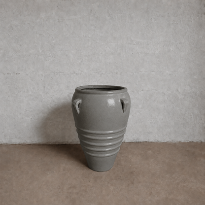 The Athens Organic Fiberglass Pot - Earth Range by Florastyle By Hingham, a large gray vase with horizontal ridges, wide rim, and two small top handles, sits on a light brown floor against a textured light gray wall.