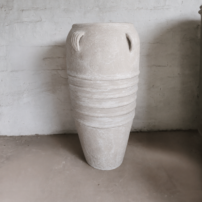 The Athens Organic Fiberglass Pot – Earth Range by Florastyle By Hingham, featuring horizontal ridges and dual handles inspired by Greek pottery, stands on a plain concrete floor against a textured, light-colored wall.