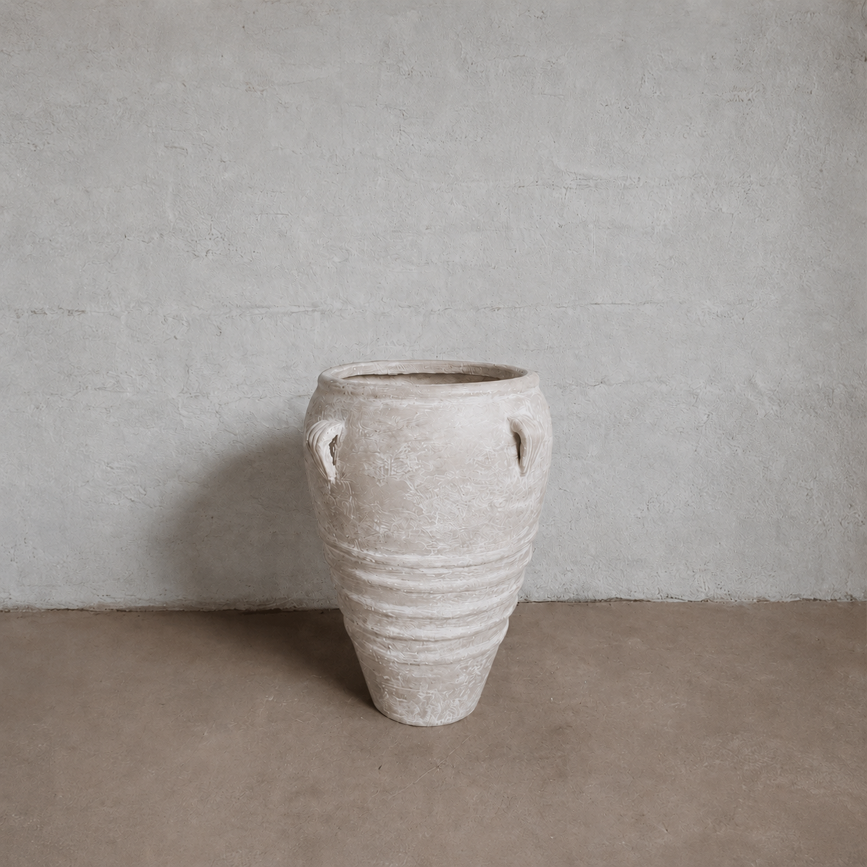 The Athens Organic Fiberglass Pot - Earth Range by Florastyle By Hingham features a large, textured beige design with two small handles, reminiscent of ancient Greek pottery, displayed on a brown floor against a light gray wall.