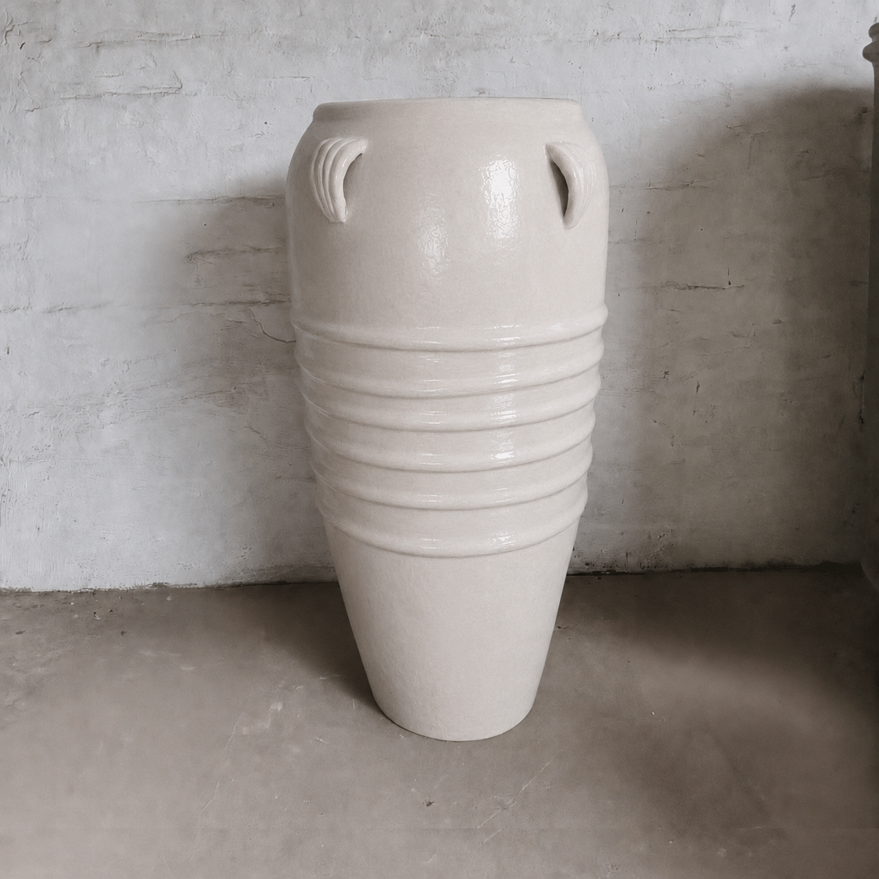 The Athens Organic Fiberglass Pot - Earth Range by Florastyle By Hingham is a large, cream vase with a tall, tapered shape, four small rim handles, and horizontal ridges, shown against a textured white wall on a concrete floor.