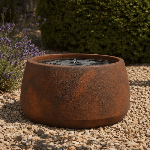 The Water Feature - Bios Low by Florastyle By Hingham is a round, textured, rust-colored piece with rippling water, placed on gravel among green shrubs and purple flowers, beautifully blending as a modern planter accent.