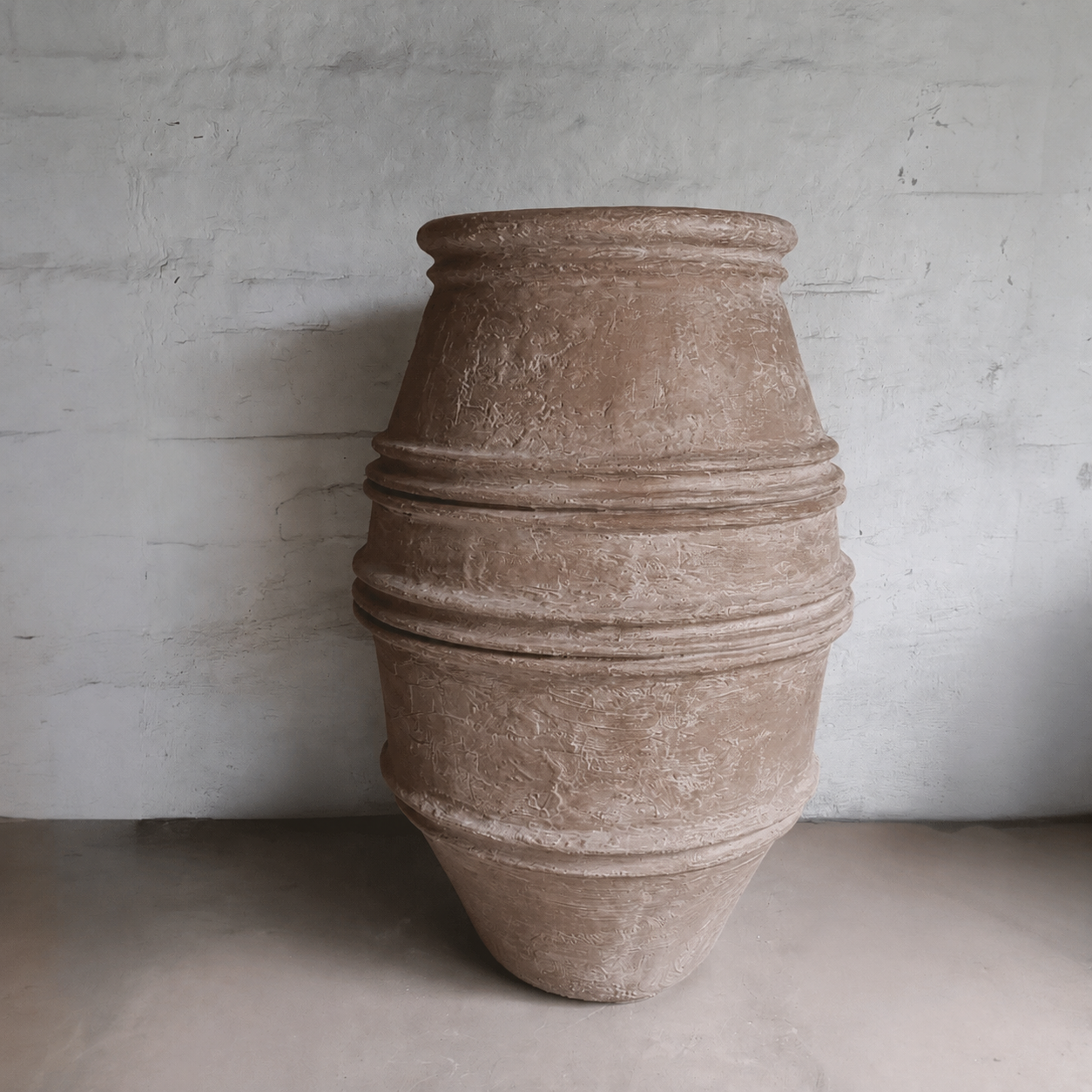 A large, textured brown Crete Organic Fiberglass Pot from Florastyle By Hingham's Earth Range stands upright against a plain, light-colored wall and floor.