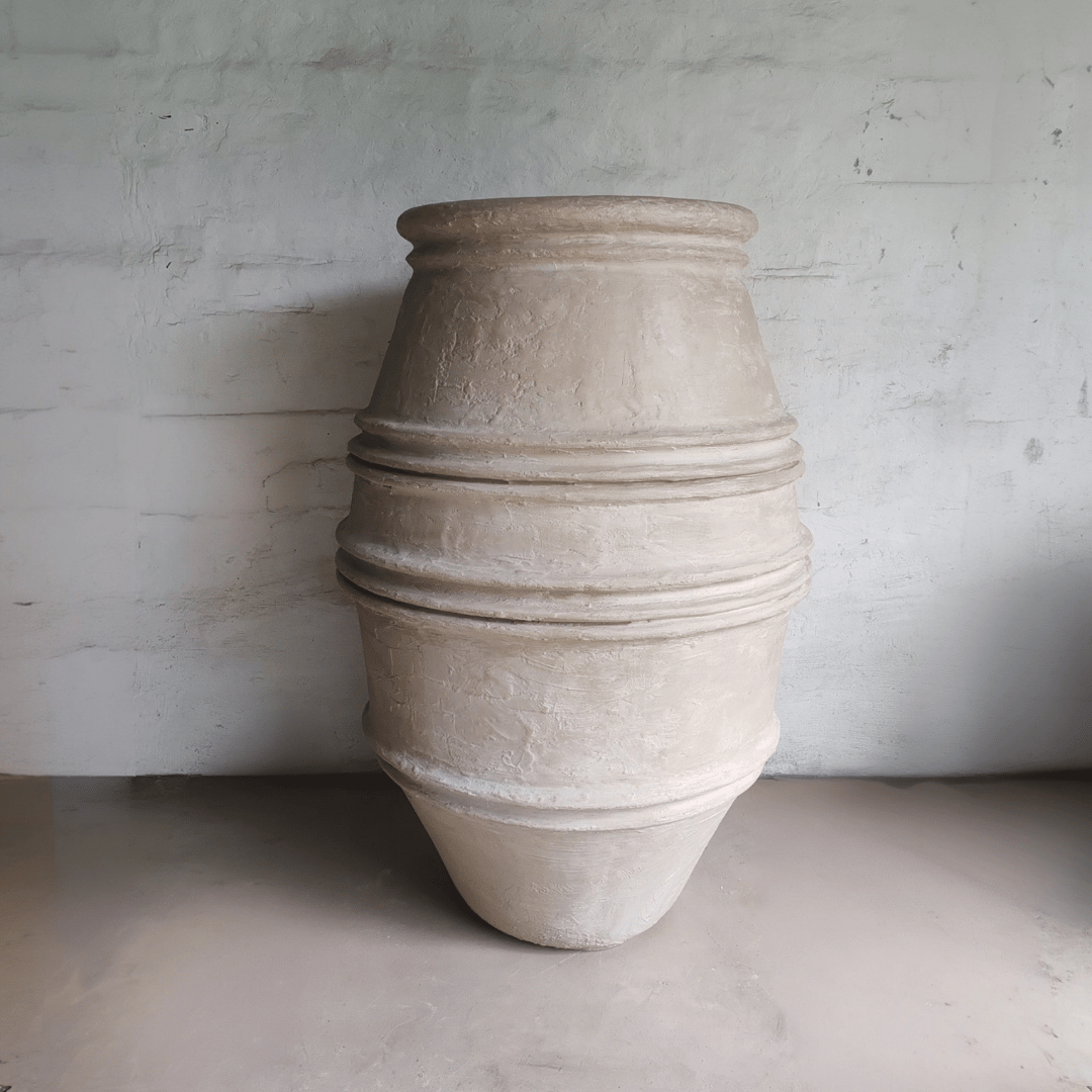 The Crete Organic Fiberglass Pot – Earth Range by Florastyle By Hingham, a large beige textured planter with three ridges, stands upright against a light gray wall—ideal as an organic modern decor accent or Mediterranean-style planter.
