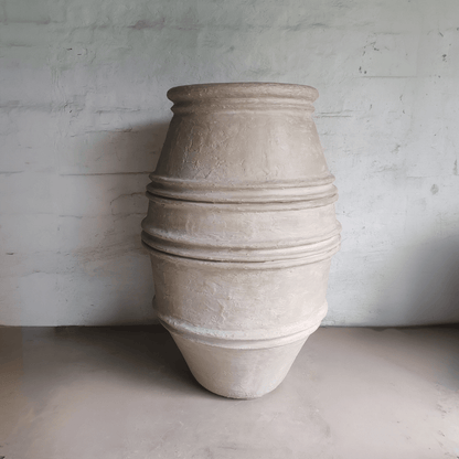 The Crete Organic Fiberglass Pot – Earth Range by Florastyle By Hingham, a large beige textured planter with three ridges, stands upright against a light gray wall—ideal as an organic modern decor accent or Mediterranean-style planter.