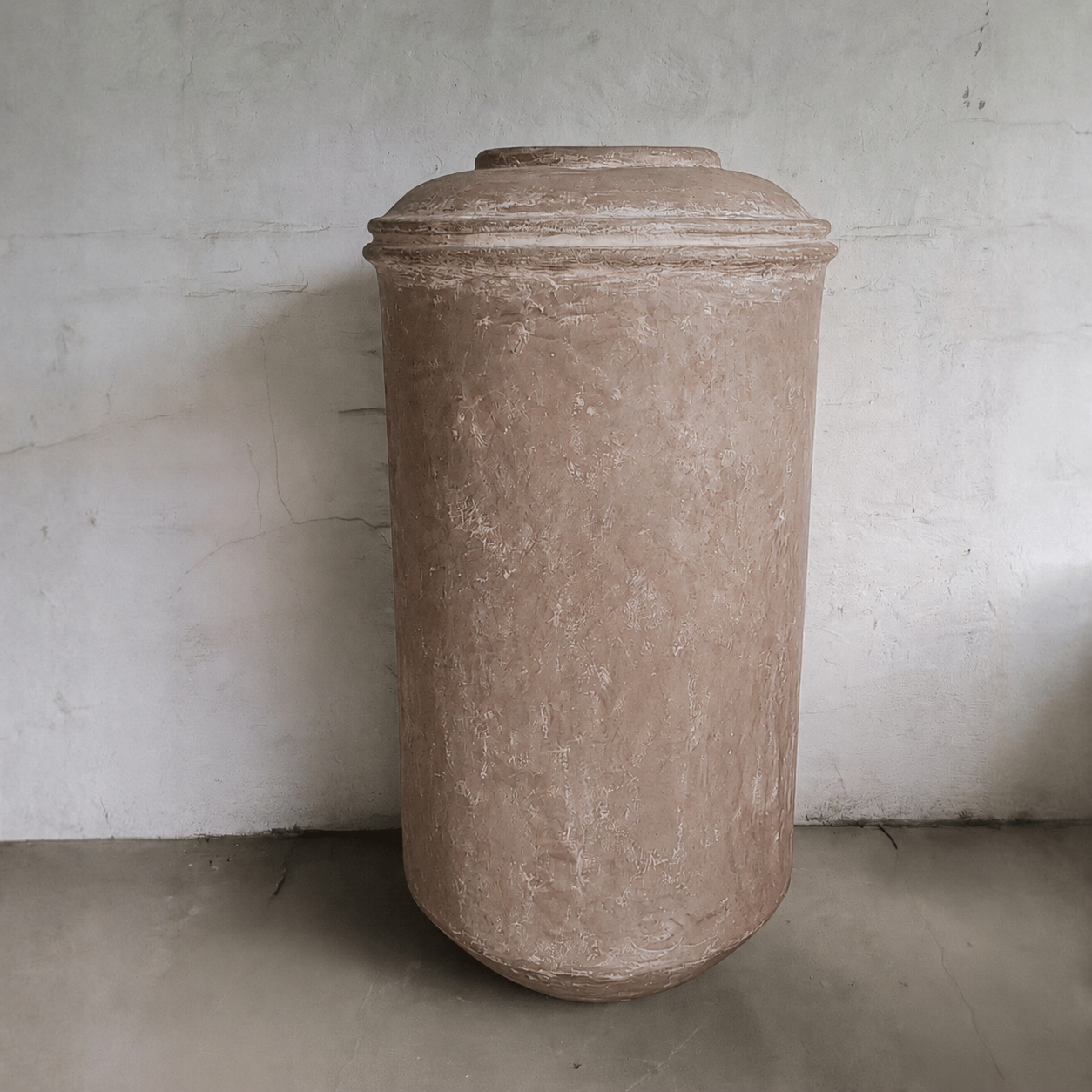 The Damascus Organic Fiberglass Pot - Earth Range by Florastyle By Hingham, a tall cylindrical planter with a rounded lid, is placed against a textured light wall on a concrete floor.