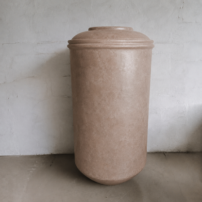 The Damascus Organic Fiberglass Pot - Earth Range by Florastyle By Hingham is a tall, cylindrical brown planter with a rounded lid, adding a modern touch to indoor spaces against light walls and concrete floors.